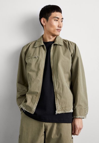 Olive green zip-up jacket with a collared neck, featuring a left chest pocket, unfinished hem, and paired with a black shirt underneath.
