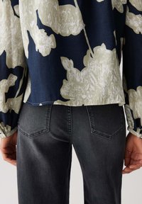 Persoon in donkergrijze jeans en een marineblauwe blouse met grote crèmekleurige bloemenprint, gezien vanaf de achterkant van de taille tot heupen.
