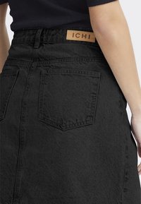 Jupe en denim noir avec une silhouette droite. Elle dispose de deux poches arrière, d'une texture subtile et d'une étiquette en cuir beige avec "ICHI" au niveau de la taille.