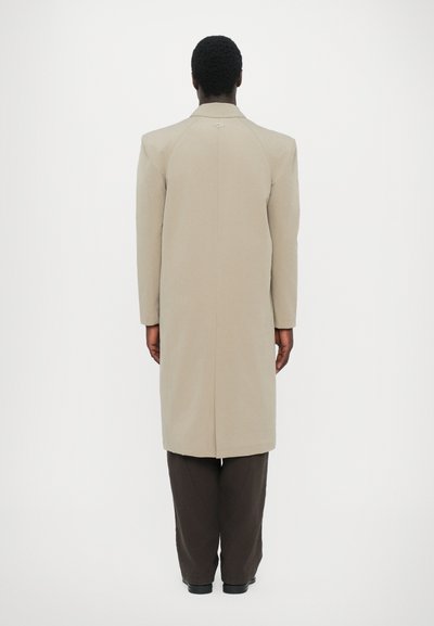Beige long coat with a structured design, single back vent, and subtle diamond accent at the collar, paired with dark brown pants.