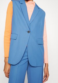 Blå ärmlös blazer med två framfickor, kragdesign och en svart knapp, tillsammans med en orange långärmad topp.