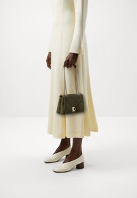 Olivově zelená prošívaná kabelka se zlatými detaily, držena v ruce, spárována s dlouhými krémovými šaty a bílými botami na blokovém podpatku.