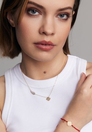 Złota biżuteria z motywem liści i kwiatów na delikatnym naszyjniku, w połączeniu z czerwonym sznurkiem bransoletki i kwiatowym akcentem.