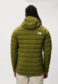 Giacca invernale imbottita di colore verde scuro con cappuccio, caratterizzata da un design trapuntato e un logo bianco sulla parte superiore della schiena. Texture liscia e forma aderente.