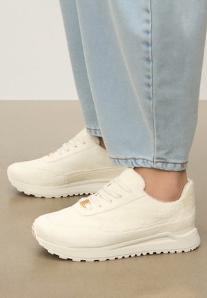Witte, harige sneakers met dikke zolen gedragen bij lichtblauwe spijkerbroeken, weergegeven op een beige vloer.