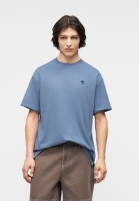 Νεαρός άνδρας που φοράει μπλε κοντομάνικη μπλούζα με μικρό λογότυπο δέντρου και καφέ παντελόνι, στέκεται μπροστά σε έναν απλό ανοιχτό γκρι φόντο.