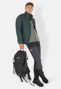 Outfit da uomo per attività all'aperto che include una giacca puffy verde scuro, un fleece grigio, pantaloni neri con tasche e uno zaino nero con una bottiglia d'acqua.