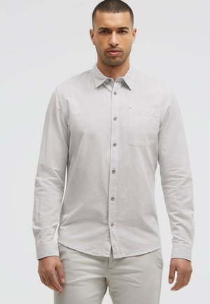 Mann mit kurzen Haaren, trägt ein hellgraues, langärmliges Hemd mit Knöpfen und hellgraue Hose, steht vor einfachem Hintergrund.