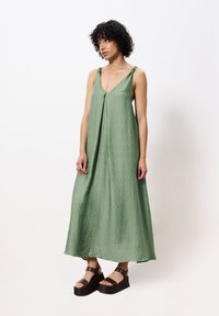 Grünes Maxikleid aus strukturiertem Material, mit tiefem V-Ausschnitt und Bindeschlaufen. Kombiniert mit klobigen braunen Plattform-Sandalen. Minimalistisches Design.