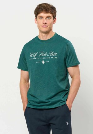 Jeune homme portant un t-shirt vert U.S. Polo Assn. et un pantalon foncé, debout les mains dans les poches devant un fond uni.