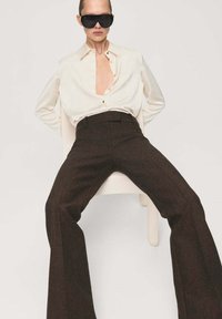 Personne portant de grandes lunettes de soleil foncées, une chemise blanche déboutonnée et un pantalon large foncé, assise les jambes écartées sur une chaise blanche.