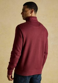 Bordeaux hoodie met hoge kraag, gestructureerde stof en reliëflogo op de kraag. Model draagt blauwe jeans en staat tegen een neutrale achtergrond.