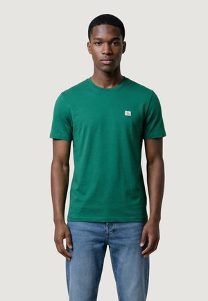Jeune homme debout, portant un t-shirt vert à manches courtes avec un petit logo sur la poitrine et un jean bleu clair, fond neutre.