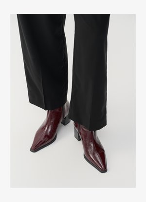 Burgundy patent leather ankle boots with a pointed toe, chunky heel, and textured finish, paired with black wide-leg trousers.