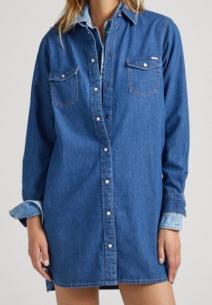 Frau trägt ein geschichtetes blaues Jeanshemdkleid mit Druckknöpfen und Brustklappen-Taschen, die Ärmel teilweise aufgerollt.