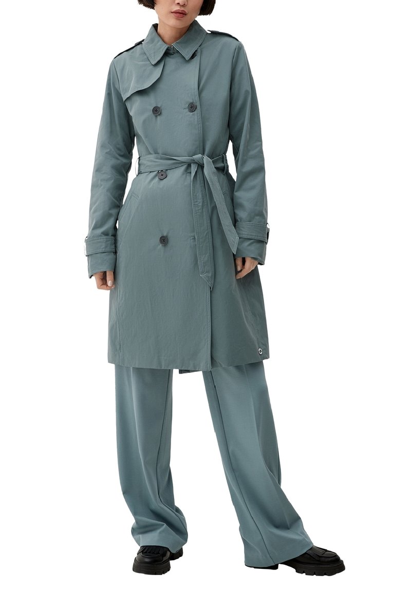 Femme portant un trench-coat bleu sarcelle à double boutonnage avec ceinture, un pantalon ample assorti et des chaussures noires, debout sur un fond blanc.
