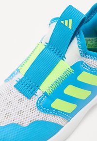 Zapatillas deportivas con parte superior de malla en blanco y azul, con detalles verdes y una correa ancha, suela texturizada y flexible.