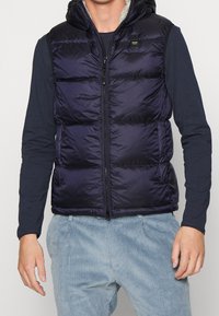 Mann trägt eine ärmellose, gepolsterte, marineblaue Weste mit Kapuze über einem langärmligen schwarzen Shirt und einer hellblauen Cordhose.