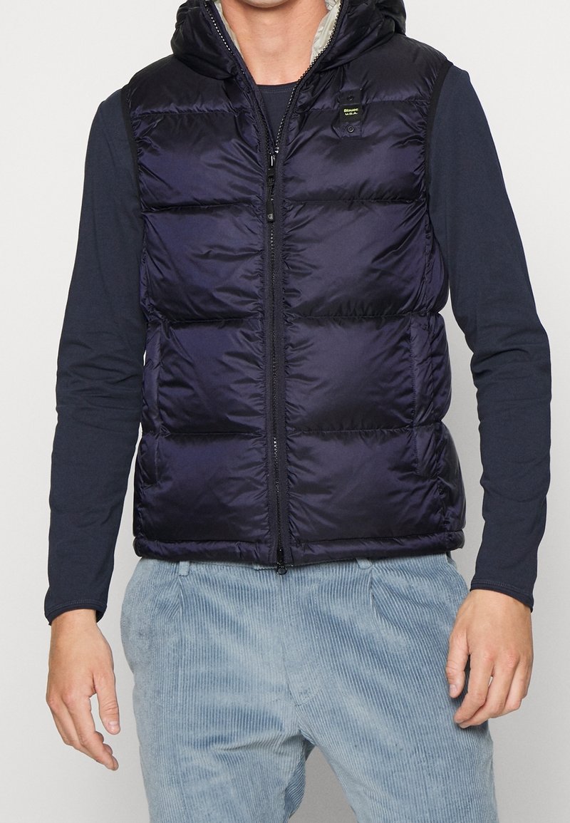 Mann trägt eine ärmellose, gepolsterte, marineblaue Weste mit Kapuze über einem langärmligen schwarzen Shirt und einer hellblauen Cordhose.