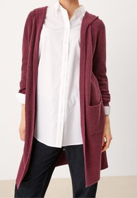 Cardigan lungo color marrone bordeaux con un motivo a maglia testurizzato, apertura frontale e due tasche anteriori, indossato sopra una camicia bianca con bottoni e jeans scuri.