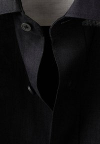 Gros plan du col d'une veste en tissu noir avec un rabat boutonné et des coutures visibles sur un fond sombre.