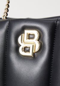 Bolsa de couro preta com um destacado logótipo em dourado e branco. Textura suave com costura visível e detalhe da alça em corrente.