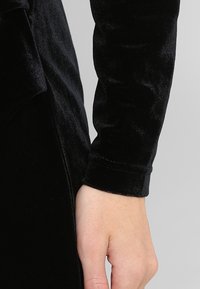 Haut à manches longues en velours noir avec une texture lisse et des poignets ajustés. Le tissu présente une finition riche et brillante.