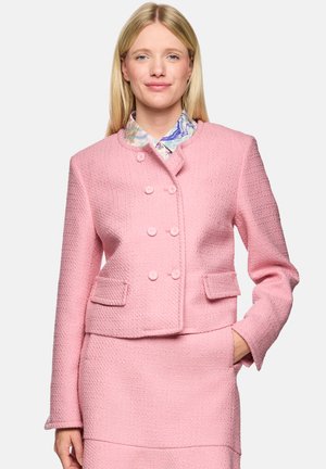 Frau trägt eine strukturierte, pinkfarbene, doppelt geknöpfte Jacke und einen passenden Rock mit einem gemusterten Hemdkragen, Hand in der Tasche, neutraler Hintergrund.