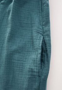 Tessuto turchese con superficie morbida e strutturata, caratterizzato da lievi pieghe e un dettaglio di tasca sul lato. Materiale liscio e leggero.