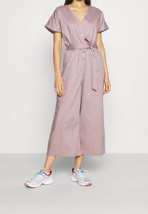 Combinaison lavande claire à manches courtes, col en V profond, boutons à l'avant, taille ceinturée et coupe jambes larges. Associée à des baskets multicolores.