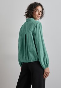 Blouse légère turquoise avec de longues manches bouffantes et un ourlet froncé. Présente un pli au dos pour un ajustement décontracté. Pantalons noirs visibles.