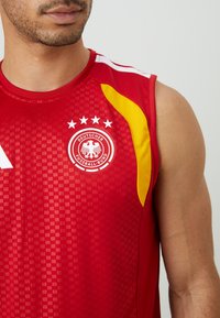 Piros sportmez, textúrált dizájnnal, sárga kiegészítővel és a "Deutscher Fussball-Bund" logóval, amely egy sast ábrázol, a logó felett négy fehér csillaggal.