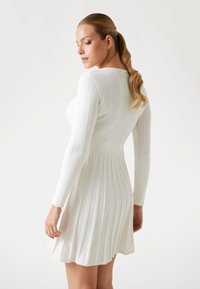Robe en tricot blanche à manches longues, présentant une texture côtelée et une jupe évasée, cintrée à la taille pour une silhouette ajustée.