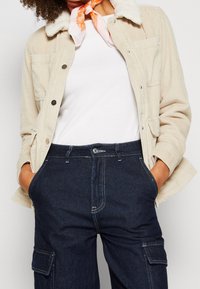 Chaqueta de pana beige con un suave collar crema, que cuenta con bolsillos frontales y botones a presión. Combinada con pantalones de cargo de mezclilla oscura.