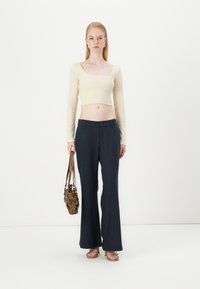 Top cropped beige a maniche lunghe abbinato a pantaloni a zampa d'elefante blu navy. Il modello tiene in mano una borsa marrone. Uno sfondo minimalista e neutro.