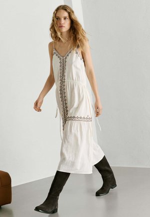 Vrouw die loopt, gekleed in een wit mouwloos midi-jurk met borduursels en zwarte kniehoge laarzen, tegen een effen lichte achtergrond.