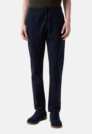 Mężczyzna ubrany w granatowe, dopasowane spodnie z ściągaczem w pasie oraz granatowe sneakersy z brązowymi podeszwami, stoi na jednolitym tle.