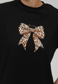 Camiseta negra con un gráfico de un lazo de estampado de leopardo en tonos de naranja y marrón. El texto sobre el lazo dice: "No seas tímido."