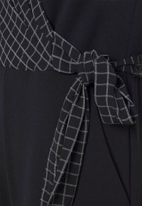 Musta mekko, jossa on ruudullinen valkoinen kuvio vyötäröllä, varustettuna sidottavalla kankaisella vyöllä. Sileä rakenne ja istuva muotoilu.