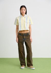 Camicia a maniche corte tagliata con strisce gialle e blu, abbinata a jeans marroni larghi e sneakers beige. Design semplice, dettaglio del logo discreto.