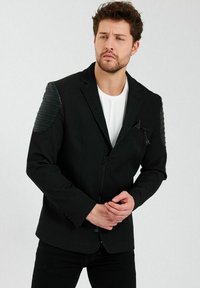 Zwarte blazer met getextureerde leren schouderaccenten, ritssluiting, notch-kraag en een getailleerd ontwerp. Gedragen over een witte shirt.