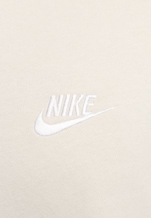 Logo de Nike sobre un fondo de tela beige claro. El logo está bordado en blanco, presentando una tipografía limpia y un diseño de swoosh.