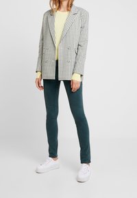 Mujer vistiendo un blazer a cuadros sobre un suéter amarillo pálido, jeans ajustados oscuros y zapatillas blancas de plataforma, de pie contra un fondo liso.