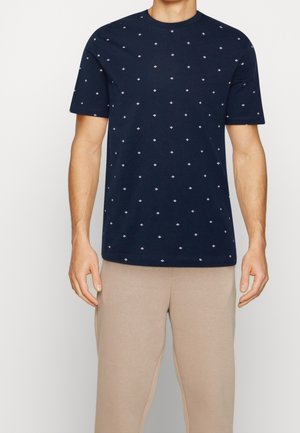 Mann trägt dunkelblaues Kurzarm-T-Shirt mit kleinem weißem Diamantmuster, kombiniert mit hellbeigen Hosen, steht vor schlichtem Hintergrund.