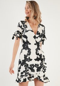 White wrap dress with black floral pattern, short ruffled sleeves, and a frilled hemline. Made of textured fabric.
