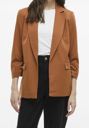 Femme portant un blazer marron avec des poches à rabat sur un haut blanc et un pantalon noir taille haute, sur un fond uni.