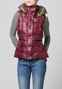 Gilet imbottito bordeaux con colletto foderato in pelliccia, caratterizzato da bottoni automatici dorati e due tasche frontali, abbinato a una maglia a maniche lunghe a righe.