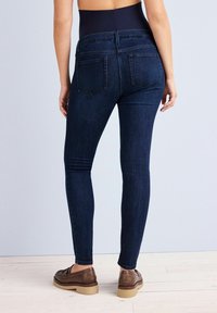 Dungarees skinny berpinggang tinggi berwarna biru tua terbuat dari denim dengan saku belakang dan jahitan samping. Dipadukan dengan sepatu loafer coklat di atas lantai kayu terang.