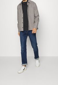 Camicia in flanella a quadri grigi con bottoni, t-shirt nera a girocollo, jeans blu e sneakers bianche con accenti neri, dotate di una vestibilità rilassata.