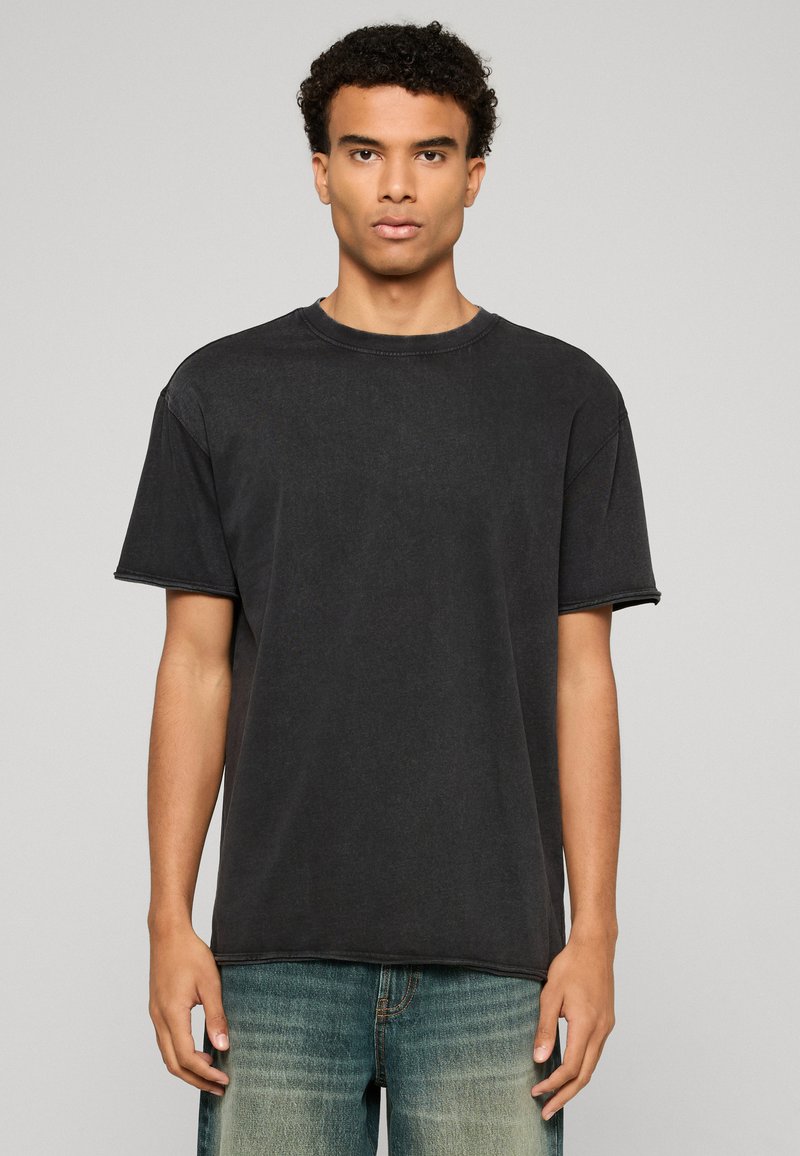 Jeune homme aux cheveux courts et bouclés portant un t-shirt noir uni à manches courtes et un jean bleu, debout devant un fond neutre.